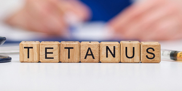 Holzwürfel mit dem Wort ‚TETANUS‘ stehen auf einer weißen Fläche, daneben liegt eine Spritze. Im unscharfen Hintergrund sind Hände und Dokumente zu sehen.